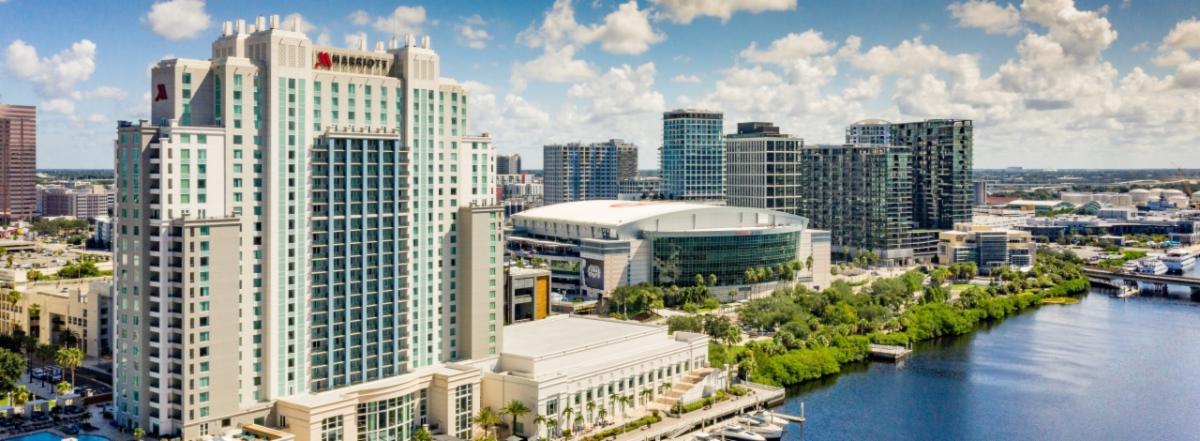 Aerial view of a waterfront city skyline with tall glass buildings and a large arena by the water.