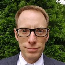 Portrait of a man with short light brown hair and black-framed glasses, wearing a dark blazer and white shirt, outdoors with greenery in the background.