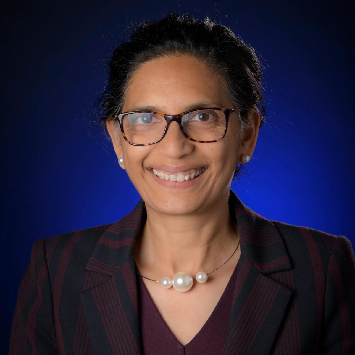 Professional headshot of a smiling woman with glasses, wearing a striped blazer and pearl necklace against a blue backdrop.