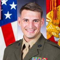 Portrait of a smiling male Marine Corps officer in dress uniform with ribbons, posed in front of the American flag (and a military flag).