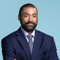 Professional portrait of a bearded man in a navy suit, light blue shirt, and patterned tie, standing against a light blue background with arms crossed.