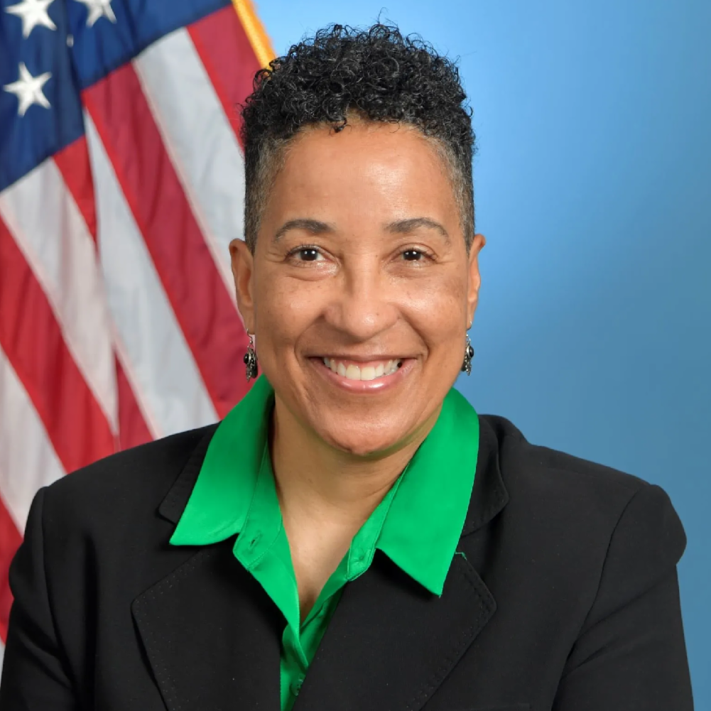 Smiling woman in a black blazer and green blouse, with the American flag in the background.