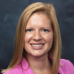 Portrait of a smiling woman with shoulder-length blonde hair, wearing a pink blazer against a dark background.