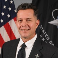 Smiling man in a dark suit with a cross lapel pin, standing in front of an American flag and government insignia background.