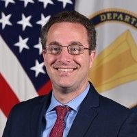 Smiling man in a dark suit with a red tie, standing in front of an American flag and a government seal backdrop.