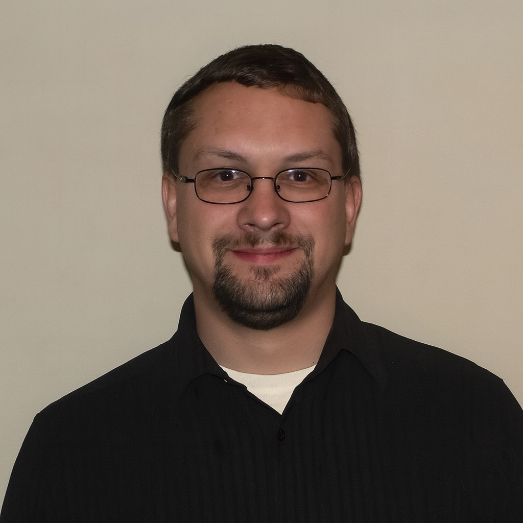 Smiling man with short dark hair and glasses, wearing a black collared shirt, posed against a beige wall.