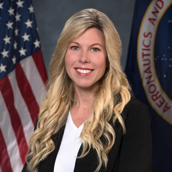Professional portrait of a smiling blonde woman in a black blazer and white top, with the U.S. flag and an agency seal in the background.