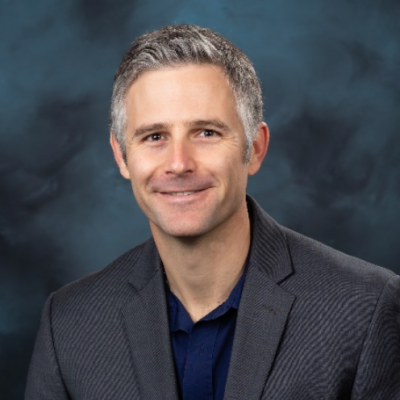 Professional head-and-shoulders portrait of a smiling man in a dark blazer and navy shirt against a blue studio backdrop.