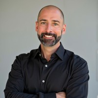 Smiling man with short hair and a beard, wearing a black button-down shirt, arms crossed against a neutral background.