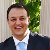 Smiling man in a dark suit and light blue tie posing outdoors with green foliage and a white column behind him.