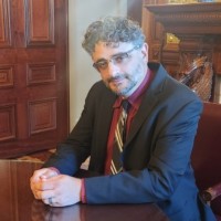 Man with gray hair and beard in a dark suit and red striped tie sits at a polished wooden desk in an office.