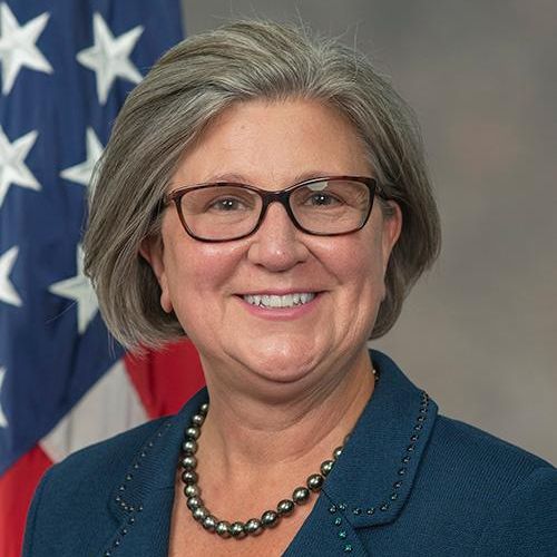 Smiling woman with short gray hair and glasses, wearing a teal blazer and pearl necklace, in front of an American flag.