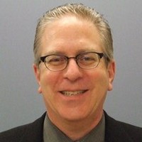 Professional headshot of a smiling middle-aged man with gray hair and glasses, wearing a dark suit and tie against a gray background.